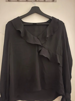 Svart blus med volang från Gina Tricot - Snygg svart blus från Gina Tricot med långa ärmar och volangdetalj längs ringningen. Blusen har en lös passform och är tillverkad i ett tunt, lätt material som ger ett elegant intryck.