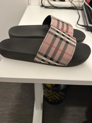 Burberry slides med rutigt mönster - Säljer ett par klassiska slides från Burberry med det ikoniska beige, svarta och röda rutmönstret på remmen. Sulan är svart och gjord i ett bekvämt syntetmaterial. Perfekta för dig som vill ha en snygg look på sommaren. Tofflorna är köpta föra sommaren och är i mycket bra skick, inga fel på tofflorna och använda bara en period, kom i dm för frågor