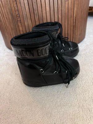 Svarta Moon Boot snow boots - Säljer ett par klassiska svarta Moon Boot snow boots med hög skaft och bred silhuett. Skorna har snörning framtill, logga runt skaftet och är tillverkade i syntetmaterial med vadderad insida. Perfekta för kalla dagar och riktigt snygga till vinterlooken.