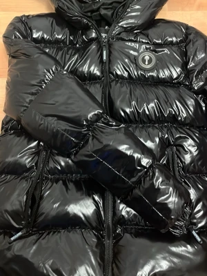 Svart glansig dunjacka från Trapstar - Säljer en svart, glansig dunjacka från Trapstar med huva och dragkedjor på fickorna. Jackan har en puffig design och logga på bröstet. Perfekt för kalla vinterdagar och riktigt snygg streetstil.