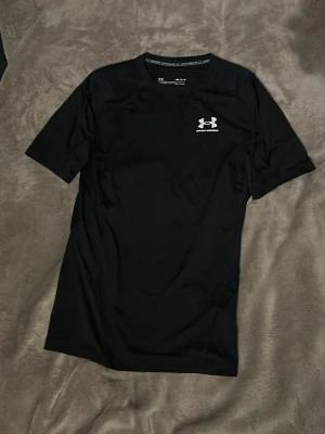 Svart träningströja från Under Armour - Säljer en svart träningströja från Under Armour med korta ärmar och rund hals. Tröjan har en diskret logga på bröstet och är tillverkad i ett lätt, snabbtorkande funktionsmaterial som passar perfekt till träning.