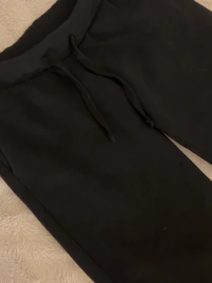 Svarta mjukisbyxor med dragsko - Ett par klassiska svarta mjukisbyxor med resår i midjan och justerbar dragsko. Byxorna har raka ben och är tillverkade i ett mjukt och bekvämt material, perfekt för chill dagar hemma eller när du vill ha det extra comfy.