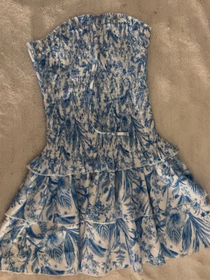 tubetopp klänning - Supersöt blåvit kortklänning med smockad överdel och volangkjol. Klänningen har ett detaljerat blommönster och är ärmlös, perfekt för varma dagar. Materialet känns lätt och luftigt, vilket gör den skön att bära.