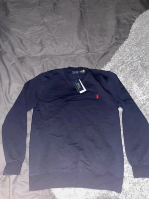 Mörkblå stickad tröja Polo Ralph Lauren - Mörkblå stickad tröja från Polo Ralph Lauren med rund halsringning och klassisk röd broderad logga på bröstet. Tröjan har långa ärmar och ribbade muddar vid ärmslut och nederkant. Perfekt för en stilren och avslappnad look.