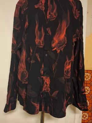 Svart skjorta med brinnande röda rosor - Unik svart skjorta med långa ärmar och knappar framtill. Skjortan har ett fett mönster av röda rosor och eldslågor som verkligen sticker ut. Perfekt för dig som vill ha något annorlunda och vågat i garderoben.