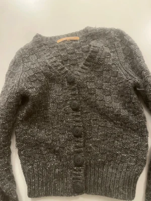 Grå glittrig kofta från Silvian Heach - Mysig grå stickad kofta från Silvian Heach med glittriga inslag och stora svarta knappar framtill. Koftan har ribbade muddar och en djup V-ringning. Perfekt för lager på lager och ger en trendig touch till din outfit.