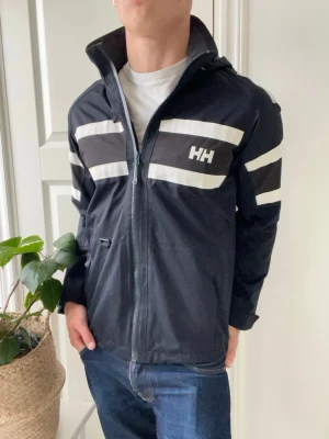 Helly Hansen regnjacka  - Helly Hansen regnjacka / vindjacka. Den klassiska helly Hansen regnjacka / vindjacka. Med HH loggan omkring jackan. Inga defekter på jackan - en väldigt trevlig jacka som passar perfekt för vintern och våren. Otroligt najs passform på modellen som är 180 cm 75 kg! 