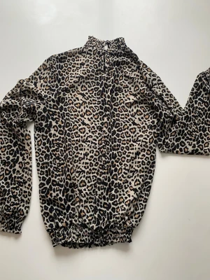 Leopardmönstrad blus med hög krage - Snygg blus med leopardmönster i beige, svart och brunt. Blusen har långärmad design, hög smockad krage och resår i både ärmslut och nederkant. Materialet känns lätt och luftigt, perfekt för att sticka ut i mängden.