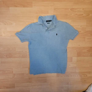 Ljusblå pikétröja från Ralph Lauren - Klassisk ljusblå pikétröja från Ralph Lauren med korta ärmar och krage. Tröjan har två knappar vid halsen och den ikoniska broderade loggan på bröstet. Perfekt till jeans eller shorts för en avslappnad men stilren look.