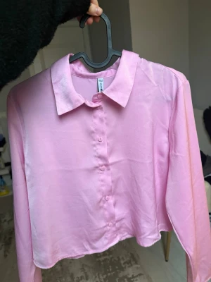 Ljusrosa croppad skjorta i satin - Trendig ljusrosa croppad skjorta med klassisk krage och knappar framtill. Skjortan har långa ärmar och är gjord i ett glansigt satinliknande material som ger en lyxig känsla. Perfekt för dig som vill sticka ut med en färgglad och modern look.