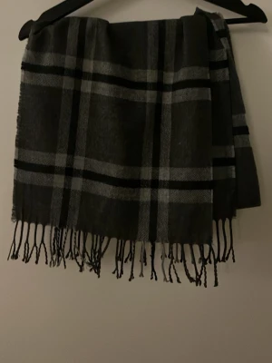 Mörkgrå halsduk med rutigt mönster - Snygg mörkgrå burberry liknandehalsduk med klassiskt rutigt mönster i svart och ljusgrått. Halsduken har fransar i ändarna och är tillverkad i ett mjukt tyg som känns skönt mot huden. Perfekt accessoar för att lyfta din outfit under kyliga dagar.