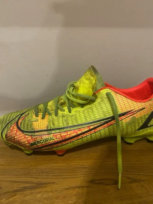 Nike Mercurial gula fotbollsskor - Säljer ett par Nike Mercurial fotbollsskor i neon-gult med röda och svarta detaljer. Storlek 43. Skorna har snörning, låg profil och mönstrad ovandel för bättre bollkontroll. Insidan är röd och yttersulan har dobbar för spel på gräs. Perfekta för dig som vill sticka ut på planen. Använda fåtal gånger på konstgräs därför lite mörkare nyanser men annars inga tecken på trasighet!👌🤩