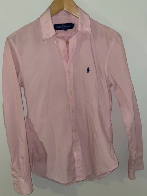 Rosa skjorta från Ralph Lauren - Klassisk rosa skjorta från Ralph Lauren med lång ärm och knappar framtill. Skjortan har den ikoniska mörkblå polospelaren broderad på bröstet och är tillverkad i ett tunt, mjukt bomullstyg. Perfekt för en stilren och avslappnad look. Det är storlek L i barn size men jag är 178 och den passar som en S skulle jag säga!