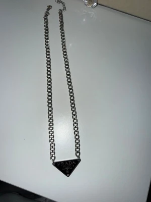 Prada silverfärgat kedjehalsband - Snyggt halsband från Prada i silverfärgad metall med en kraftig kedja och en ikonisk triangelformad platta med logotypen 'PRADA MILANO' framtill. Perfekt statement piece för dig som gillar exklusiva accessoarer och vill sticka ut. Ej riktig 