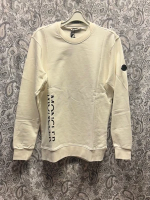 Vit sweatshirt från Moncler - Snygg vit sweatshirt från Moncler med svart logotyptryck längs sidan och en rund halsringning. Tröjan har långa ärmar, ribbade muddar och en diskret Moncler-patch på ärmen. Tillverkad i mjukt bomullsmaterial för en clean och stilren look.