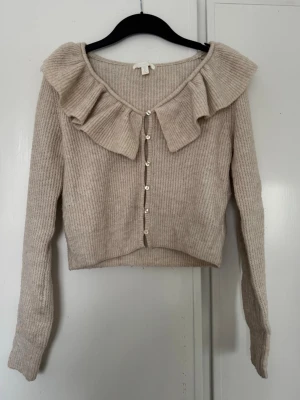 Beige kofta med volangkrage - Supersöt beige ribbstickad kofta med volangkrage och pärlknappar framtill. Kortare modell med långa ärmar och mjukt material. 