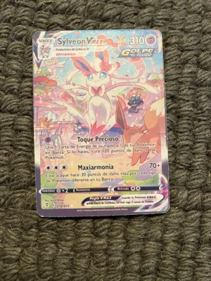 Sylveon VMAX Pokémonkort - Unik Sylveon VMAX Pokémonkort med färgstark och glittrig design. Kortet har motiv av Sylveon i pastellfärger och är på spanska. Perfekt för samlare eller Pokémonfans som vill utöka sin samling med ett VMAX-kort.