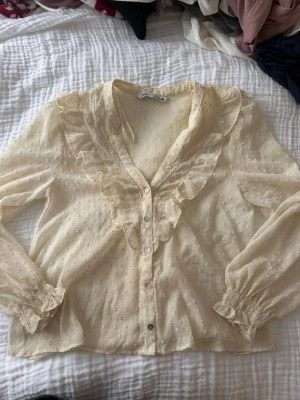 Krämvit blus med volanger från Zara - Söt krämvit blus från Zara med volangkant längs knäppningen och v-ringning. Blusen har diskreta prickar i tyget, transparenta långärmar med resår vid ärmslut och fina spetsdetaljer. Perfekt för en romantisk look.