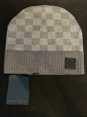 Grå mössa - Snygg grå mössa från med diskret rutmönster och ribbad kant. Märkt med en svart patch framtill och klassisk tag. Tillverkad i mjuk ullmix som håller dig varm och stilren under kalla dagar.