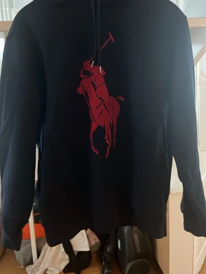 Marinblå hoodie från Polo Ralph Lauren - Snygg marinblå hoodie från Polo Ralph Lauren med stor röd polospelare tryckt på bröstet. Klassisk huva med dragsko och långa ärmar. Perfekt för en avslappnad eller festlig outfit.