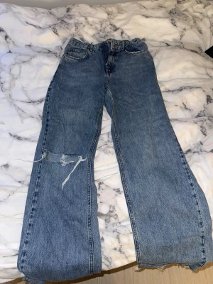 Blåa raka jeans från Gina tricot - Säljer ett par blåa raka jeans med hål på vänster ben. Jeansen har klassisk femficksdesign och hög midja. Perfekta för en avslappnad och trendig look.