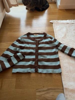 Randig brun och ljusblå kofta - Snygg ribbstickad kofta med breda bruna och ljusblå ränder. Kortare modell med rund halsringning och knappar framtill. Perfekt för att addera lite färg till din outfit.