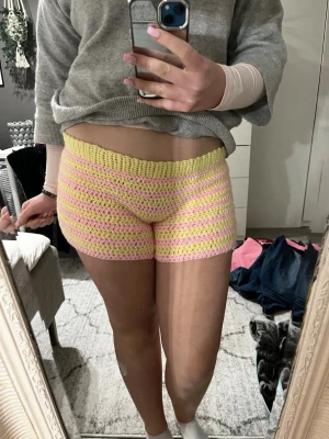 Virkade shorts i pastellrosa och gult - Säljer ett par handvirkade shorts i pastellrosa och gult med randigt mönster. De är låga i midjan och har en tight passform. Perfekta för dig som vill ha något unikt och färgglatt i garderoben då de är egen gjorda!
