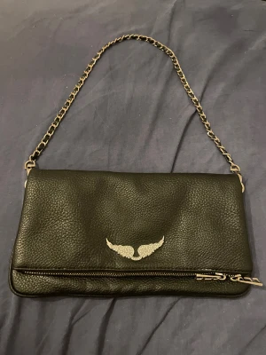 Zadig & voltaire - Snygg svart axelväska i skinnimitation med silverfärgad kedjerem och dragkedjedetaljer. Väskan har ett coolt vingmotiv i silver framtill och praktiska fack med dragkedja både fram och bak. Perfekt för dig som gillar stilrena accessoarer med en twist. Själv loggan sitter lite löst men märks inte om man inte pillar, går o lösa med lite limm