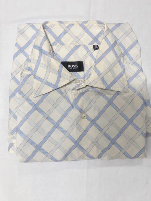 Rutig skjorta från BOSS i bomull - Snygg rutig skjorta från BOSS i vitt och ljusblått. Skjortan har klassisk krage, långa ärmar och knäppning framtill. Tillverkad i mjuk bomull som känns skön mot huden. Perfekt för dig som gillar stilrena och fräscha mönster. Bra skick.