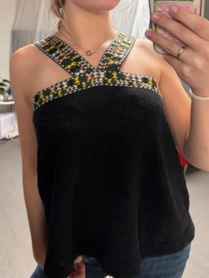 Svart linnetopp med mönstrade band - Säljer en svart linnetopp med breda axelband i färgglatt geometriskt mönster i gult, orange, grönt, svart, rosa och vitt. Toppen har korsade band framtill och är ärmlös med loose passform. Perfekt för dig som vill ha en unik och bekväm topp till sommaren.