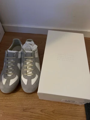 Maison margiela  - Snygga sneakers i vitt och ljusgrått med ovandel i mocka och skinn. Klassisk design med snörning och platt sula i gummi. Perfekta för dig som gillar stilrena och tidlösa sneakers. Märket Maison Margiela väldigt fina o sköna skor passar perfekt inför kommande årstider våren och sommaren väldigt stiliga och passar med allt. PRISET KAN DISKUTERAS:) Kvitto finns!!