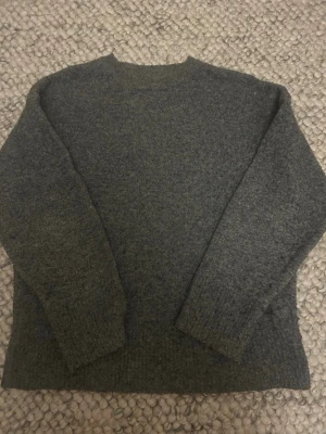 Mörkgrå stickad tröja Primark - Mysig mörkgrå stickad tröja från Primark med enkel design och rund hals. Perfekt för lager på lager och passar till allt. Tröjan har långa ärmar och är gjord i ett mjukt material som håller dig varm under kyliga dagar.