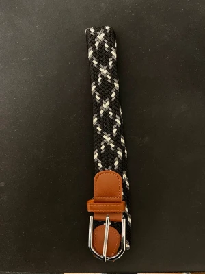 flätat grisch bälte - Snyggt flätat bälte i svart och vitt med bruna skinn-detaljer och silverfärgat spänne. Perfekt accessoar för att lyfta din outfit och passar till både jeans och chinos. Bältet har ett randigt mönster och är gjort i syntetmaterial med detaljer i skinn. Knappt använd