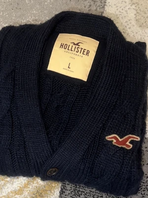 Mörkblå kabelstickad kofta Hollister - Snygg mörkblå kabelstickad kofta från Hollister med knappar framtill och två fickor. Koftan har ribbade muddar, V-ringning och det klassiska Hollister-logotypen broderad på bröstet. Perfekt för lager på lager och en chill look.