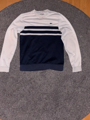 Vit och marinblå sweatshirt Lacoste - Snygg sweatshirt från Lacoste i vitt och marinblått med två mörkblå ränder över bröstet. Klassisk rund halsringning och lång ärm. Ikoniska Lacoste-loggan broderad på bröstet. Perfekt för en sportig och stilren look.