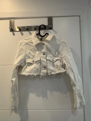 Vit croppad jeansjacka - Snygg vit croppad jeansjacka med rå kant nertill och klassiska bröstfickor med lock. Jackan har långa ärmar och knäppning framtill. Perfekt för dig som vill ha en trendig och avslappnad look.