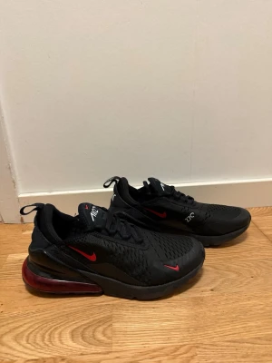 Nike Air Max 270 svarta/röda sneakers - Nike Air Max 270 sneakers i svart mesh med röda detaljer och synlig Air Max-dämpning i hälen. Snygg design med röda swoosh-loggor och 270-tryck på sidan. Skorna har en sportig look och är perfekta för dig som gillar streetstyle.