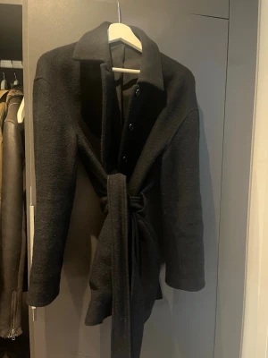 Svart Filippa K kappa - Stilren svart kappa med klassisk krage, knappar framtill och knytskärp i midjan. Kappan har en rak passform och är tillverkad i ett mjukt, ull-liknande material. Perfekt för kyliga dagar och ger en snygg siluett.nypris 4100kr 