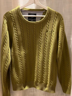 Olivgrön stickad tröja med armbågslappar - Säljer en olivgrön stickad tröja från Bondelid  med klassiskt flätmönster och rund hals. Tröjan har ribbade muddar och bruna armbågslappar i mockaimitation som ger en snygg kontrast. Perfekt för dig som gillar stilrena detaljer.