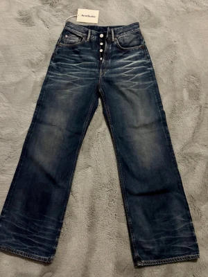 Acne Studios raka blå jeans Ny med etikett, EU 26/32 - Säljer ett par raka blå jeans från Acne Studios med snygg slitning och knappar i gylfen. Jeansen har klassisk femficksmodell och är tillverkade i kraftigt denimtyg. Perfekta för en avslappnad och trendig look.