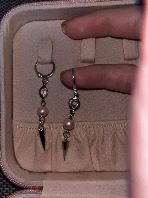 Silvriga örhängen med pärlor och spikes - Säljer ett par unika silvriga örhängen med små ljusrosa pärlor och coola metallspikar som hänger längst ner. Örhängena har en modern design med ringfäste och detaljer i metall. Perfekta för dig som vill sticka ut med din stil.