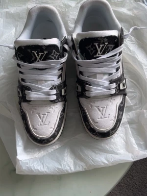 Louis Vuitton monogram sneakers - Säljer ett par snygga Louis Vuitton sneakers i vitt och svart med klassiskt LV-monogram. Skorna har vita snören, präglad LV-logga på tån och detaljer i svart läder med monogrammönster. Perfekta för dig som vill sticka ut med exklusiva sneakers.