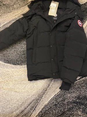 Svart dunjacka från Canada Goose - Säljer en svart dunjacka från Canada Goose med klassisk patch på ärmen och dragkedja samt knappar framtill. Jackan har huva och är tillverkad i slitstarkt material med dunfyllning som håller dig varm under kalla vinterdagar.
