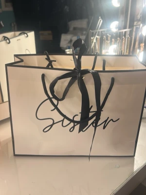 Vit presentpåse med svart text - Stilren vit presentpåse med svart text där det står 'Sister' på framsidan. Påsen har svarta band som handtag och en dekorativ rosett framtill. Perfekt för att ge bort presenter eller använda som accessoar.