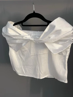 Vit axelbandslös topp med stor rosett - Supersnygg vit axelbandslös topp med en stor rosett framtill. Toppen är croppad och har en elegant offshoulder-design som verkligen sticker ut. Perfekt för dig som vill ha något unikt och trendigt i garderoben.