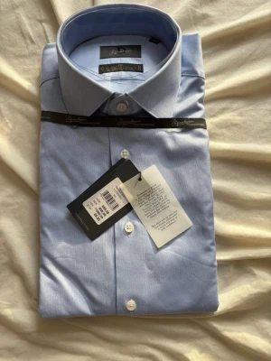 Ljusblå skjorta från Dressmann Signature - Klassisk ljusblå skjorta från Dressmann Signature, tillverkad i exklusivt bomullstyg från Tessitura Monti. Skjortan har vit knappstängning och en stilren krage, perfekt för en clean och snygg look.