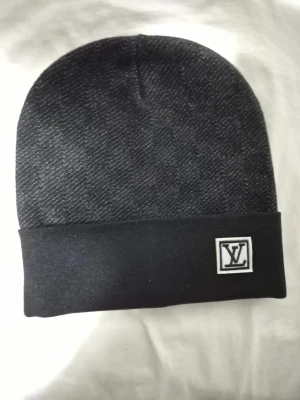 Svart mössa från Louis Vuitton - Snygg svart mössa från Louis Vuitton med diskret LV-logga framtill. Mössan har en uppvikt kant och är tillverkad i ett mjukt, stretchigt material som sitter skönt på huvudet. Perfekt accessoar för att lyfta din streetwear-look.