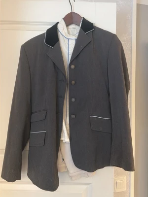 Komplett tävlingsset till barn - Säljer en klassisk mörkgrå ridkavaj med svart sammet på kragen och vita detaljer, en vit tävlings skjorta med ljusblåa detaljer. Kavajen och skjortan är från jacson wear. Säljer även ett par vita ridbyxor från Domani med förstärkning vid knäna och dragkedjeficka. Perfekt för tävling inom ridsport. Kavaj och skjorta är i storlek 150 och byxorna är i 152