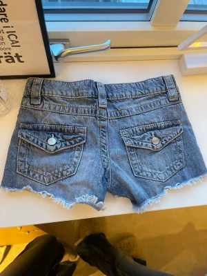 Blå jeansshorts med fransig kant - Säljer ett par blå jeansshorts med fransig kant och slitna detaljer. Shortsen har fem fickor, bälteshällor och knäppning fram. Perfekta för varma dagar och en avslappnad stil.152 