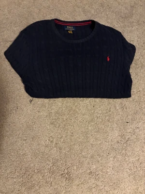 Mörkblå kabelstickad tröja Polo Ralph Lauren - Kabelstickad mörkblå tröja från Polo Ralph Lauren med rund halsringning och röd broderad logga på bröstet. Tröjan har långa ärmar och ribbade muddar vid ärmslut och nederkant. Klassisk och tidlös design som passar till det mesta. Den är i storlek xl för barn som motsvarar en S eller till och med M. Den är endast använd ett fåtal gånger.